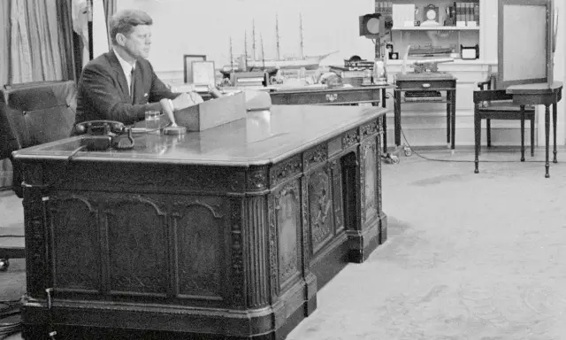John F. Kennedy am Schreibtisch im Oval Office. Hinter ihm ein Modellschiff, das an die HMS Resolute erinnert, die dem Schreibtisch seinen Namen gab.