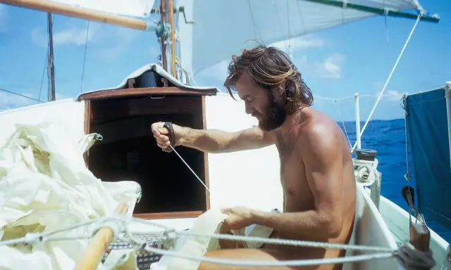 Wilfried Erdmann, mit Bart und langen Haaren, arbeitet an Bord einer Segelyacht. Er zieht an einer Leine, während die Segel im Wind wehen.