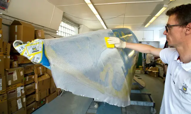 Mann bearbeitet mit gelbem Werkzeug und Handschuh ein Bootsteil. Epoxidharz Bootsbau-Material wird aufgetragen.