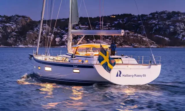 Eine Hallberg-Rassy 69 gleitet bei Dämmerung über das Wasser, die schwedische Flagge weht im Wind.