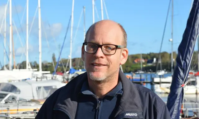 Mann mit Brille lächelt vor Segelbooten. Henningsen und Steckmest sind bereit für die nächste Ausfahrt.