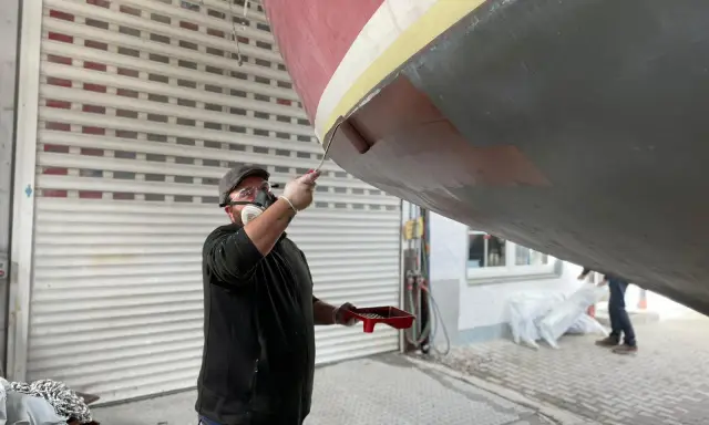 Mann mit Maske und Schutzbrille streicht mit einer Rolle den Rumpf eines Bootes.