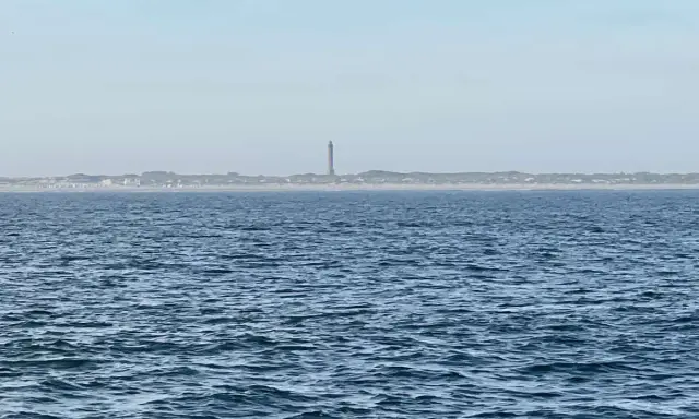 Der Leuchtturm Norderney ragt über die Dünenlandschaft am Horizont. Das weite Meer liegt davor.