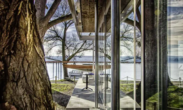 Ein Boot liegt aufgebockt am Ufer, daneben ein Tisch und Stühle. Die moderne Architektur des **Keller Werft**-Gebäudes spiegelt sich im Wasser.