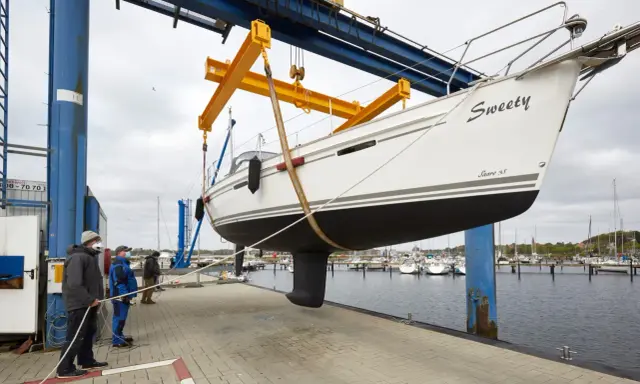 Segelboot "Sweety" wird für das unterwasserschiff reinigen aus dem Wasser gehoben. Männer sichern das Boot mit Seilen.