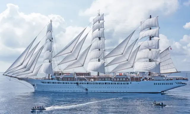 Ein majestätisches Segelschiff mit vollen weißen Segeln, das wie ein **sea cloud spirit** über das blaue Meer gleitet. Kleine Beiboote sind in der Nähe.