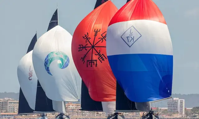 Segelboote der j class rasen über das Meer. Bunte Spinnaker mit Logos und Symbolen, im Hintergrund eine Küstenstadt.
