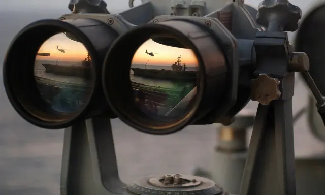 Fernglas-Werte: Ein Flugzeugträger und ein Hubschrauber spiegeln sich im Okular eines Fernglases bei Sonnenuntergang.