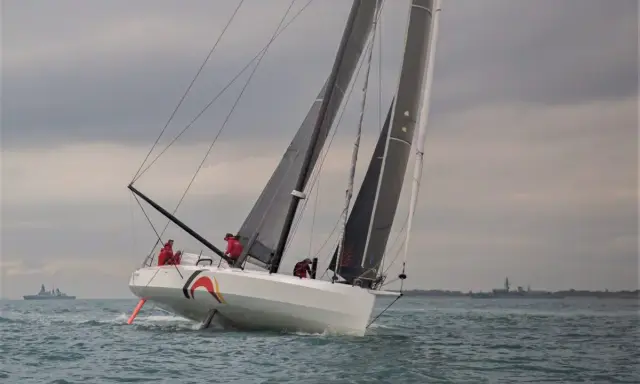 Ein IMOCA 60 segelt auf stürmischer See, die Crew in roten Jacken bereitet sich auf die nächste Welle vor.