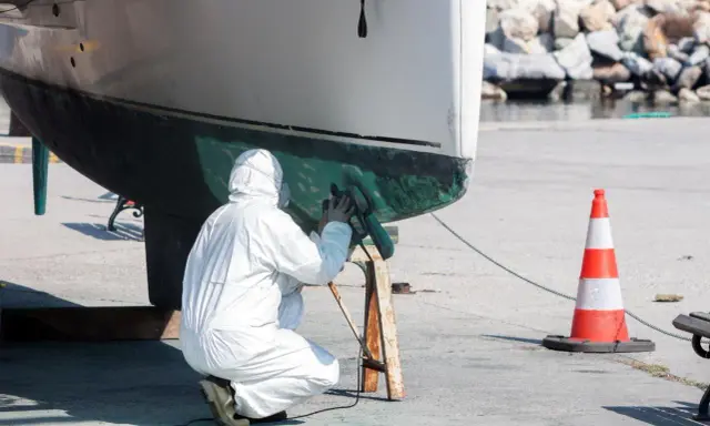 Person in Schutzanzug schleift den Rumpf eines Bootes. Die Arbeit am Boot schleifen erfordert Sorgfalt.