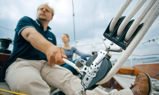 Mann zieht mit kräftiger Hand das Tauwerk am Segel. Ein Mann und eine Frau auf einem Segelboot, die das Tauwerk am Segel bedienen.