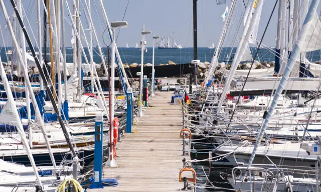 Viele Segelboote liegen im **Flensburg Hafen** an einem Holzsteg. Im Hintergrund segeln große Schiffe auf dem Meer.