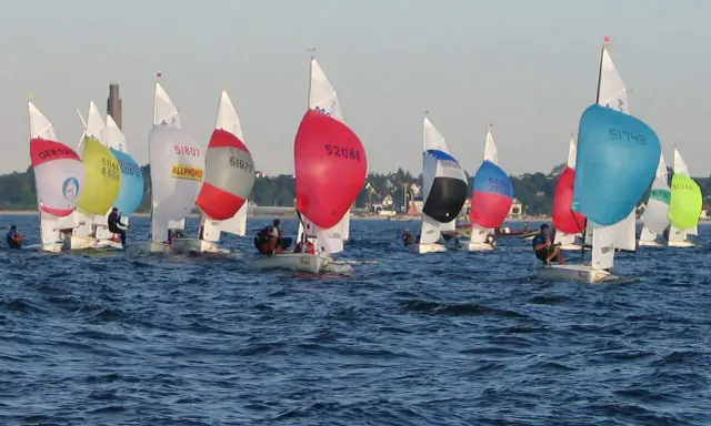 Bunte Segel dominieren das Bild einer spannenden Regatta. Mehrere kleine Boote mit Seglern sind auf dem Wasser unterwegs.