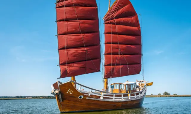 Ein hölzernes Dschunke Schiff mit roten Segeln gleitet auf ruhigem Wasser unter blauem Himmel.