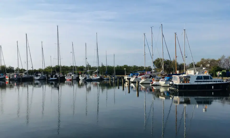 Segelboote und Motorboote liegen friedlich im Hafen Neuendorf, ihre Spiegelbilder tanzen auf dem ruhigen Wasser.