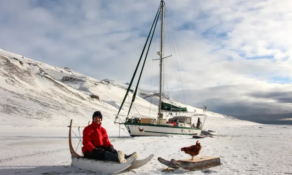 Guirec Soudée sitzt in einem Schlitten vor seinem Segelboot in einer verschneiten Landschaft. Eine Henne steht auf einem Holzbrett daneben.