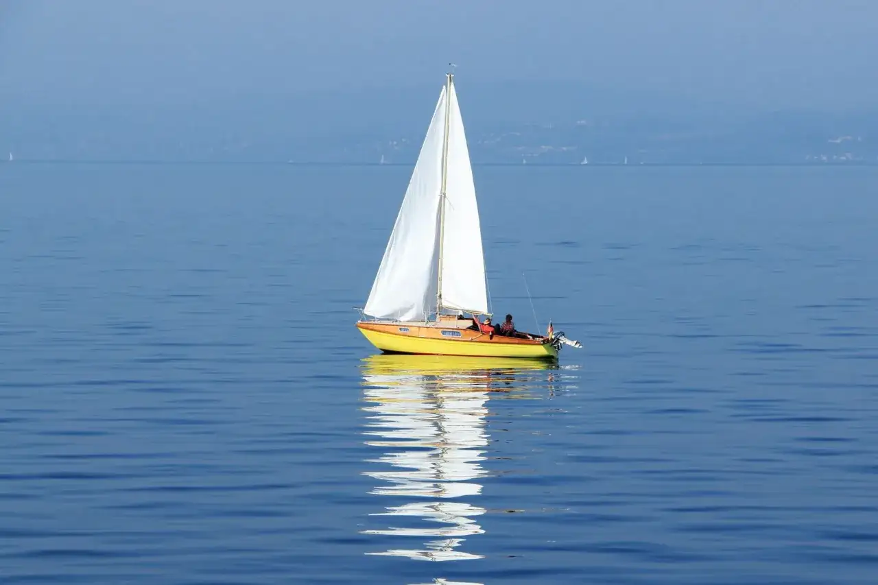 Ein gelbes Segelboot mit wei&szlig;em Segel gleitet auf ruhigem Wasser. Im Hintergrund sind weitere kleine Segelboote und eine ferne K&uuml;stenlinie zu sehen, die verschiedene kleine Segelboote Typen andeuten.