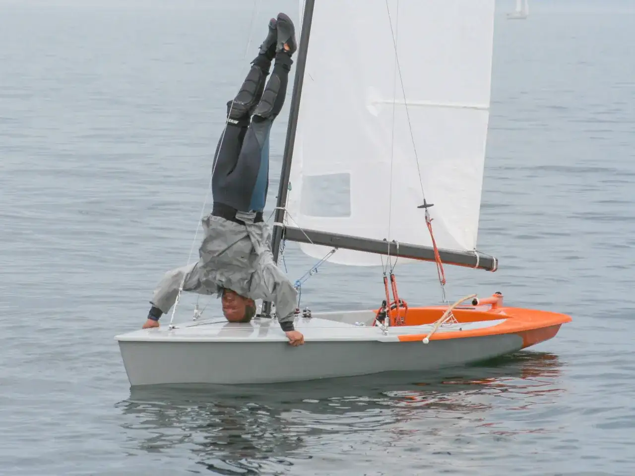 Ein Mann macht einen Handstand auf einer kleinen Jolle. Was ist eine Jolle? Ein kleines Segelboot, perfekt für waghalsige Manöver!