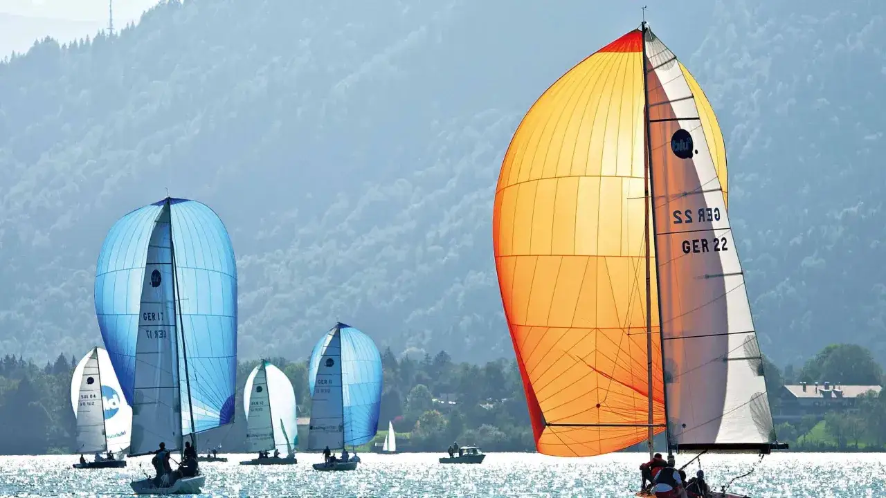 Segelboote mit bunten Spinnakern auf dem Wasser. Ein orangefarbenes Segel dominiert, w&auml;hrend im Hintergrund blaue Segel und Berge zu sehen sind. Perfekt f&uuml;r einen Podcast &uuml;ber Segeln.