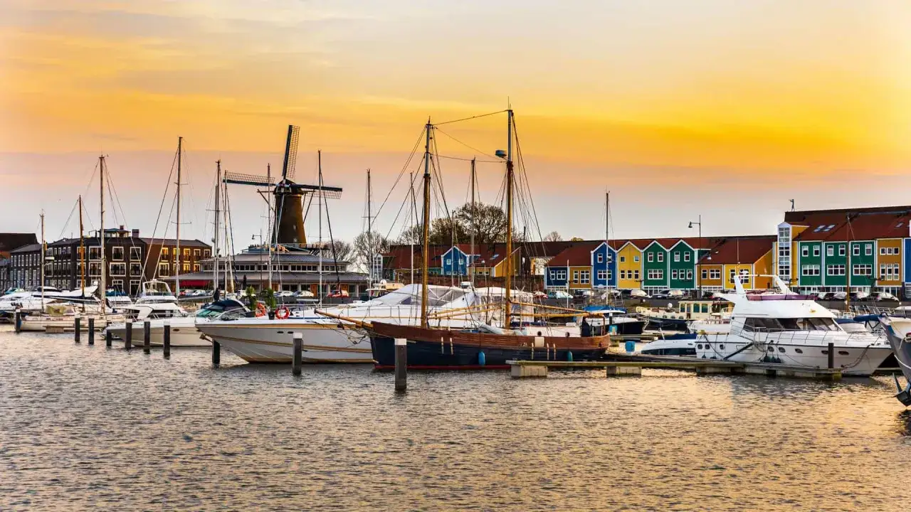 Bunte H&auml;user und ein historischer Windm&uuml;hle s&auml;umen den lelystad hafen, wo Yachten und Segelboote im goldenen Abendlicht ruhen.