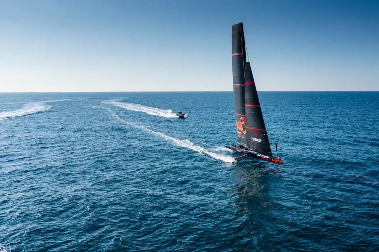 Alinghi Red Bull Racing segelt mit voller Kraft über das blaue Meer. Ein Begleitboot folgt der Spur.