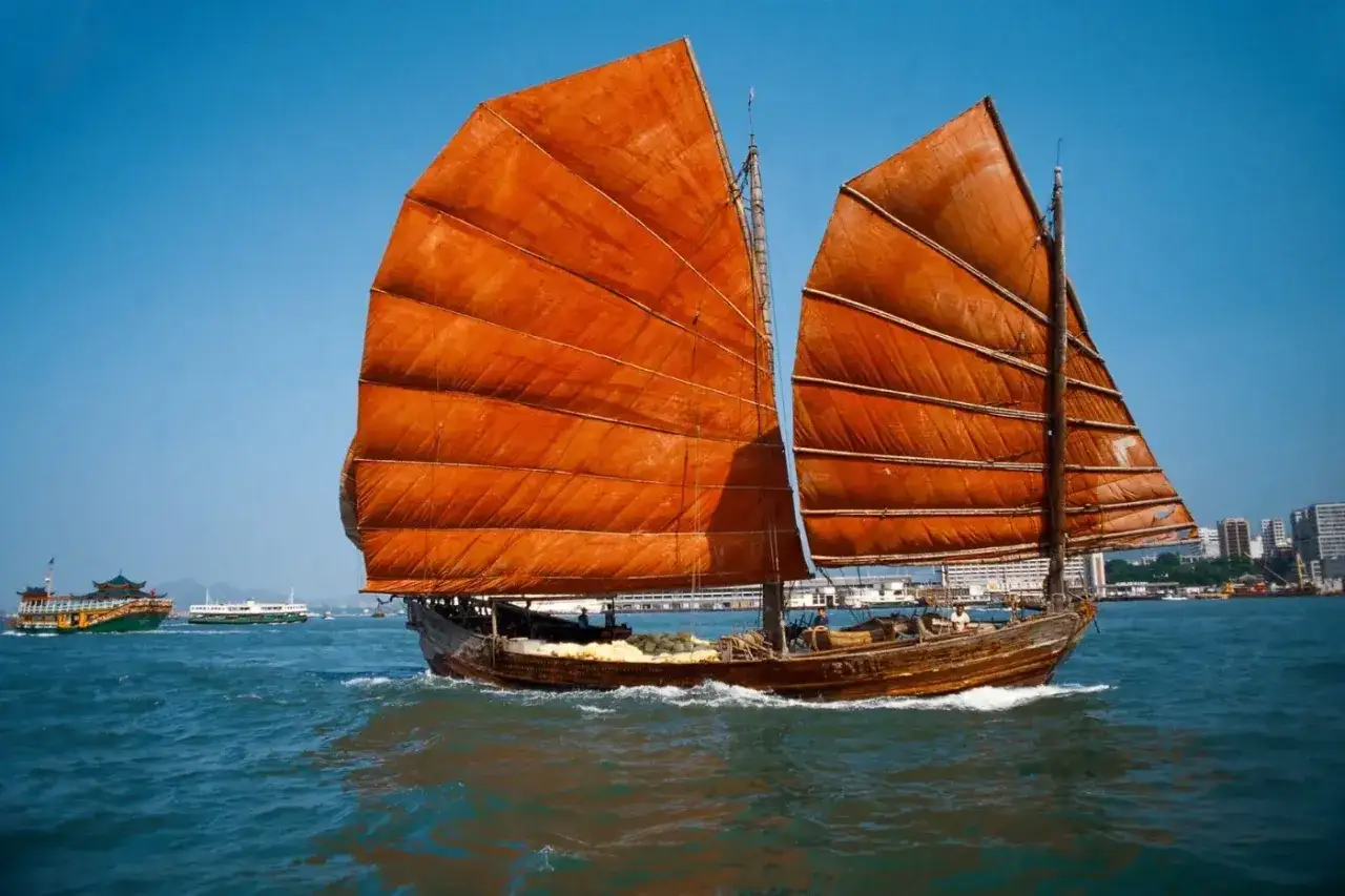 Ein traditionelles dschunke schiff mit orangefarbenen Segeln gleitet über das Meer. Im Hintergrund sind weitere Schiffe und eine Stadt zu sehen.