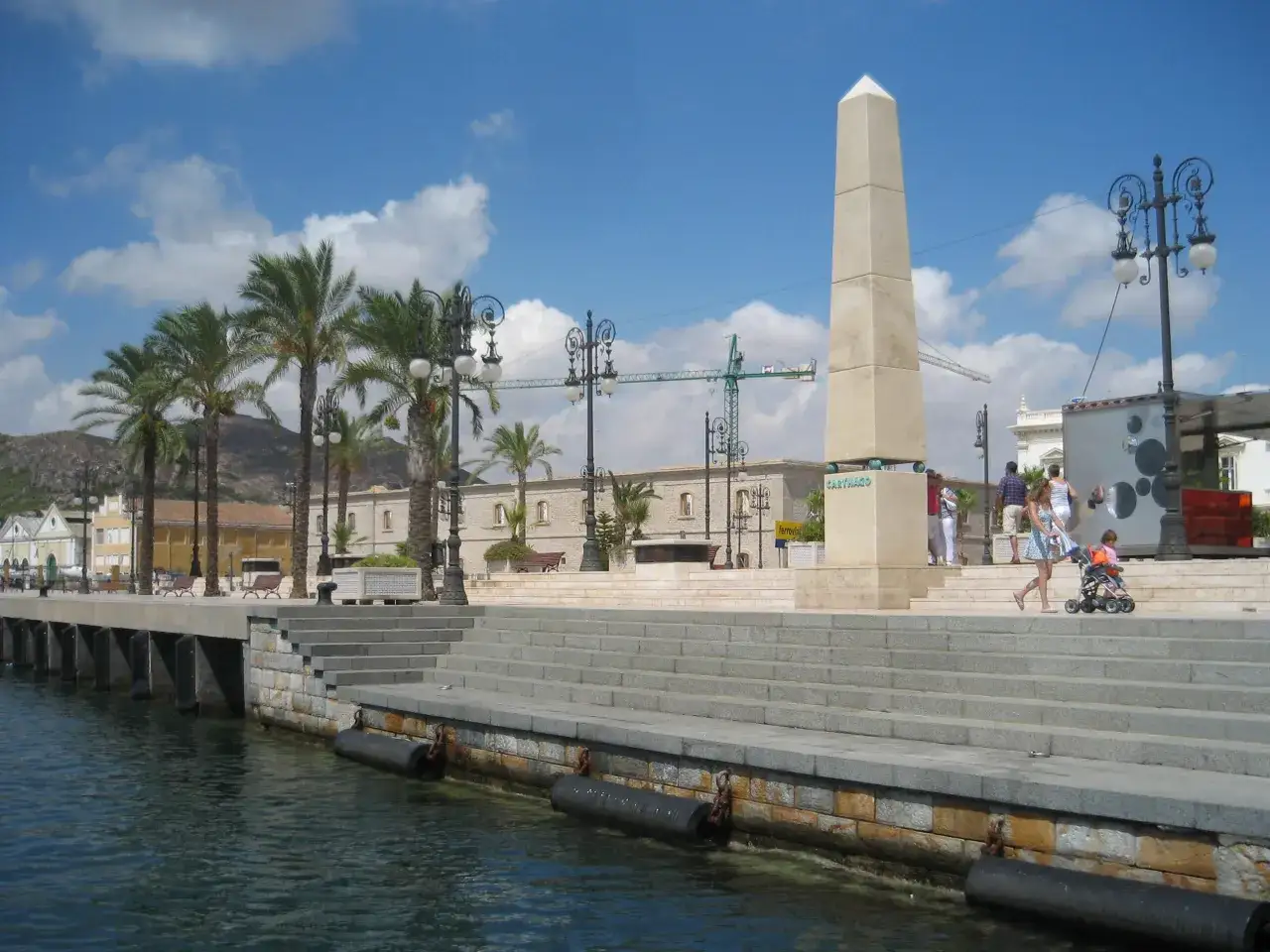 Ein sonniger Tag im Cartagena Hafen. Palmen säumen die Promenade, ein Obelisk ragt in den blauen Himmel.