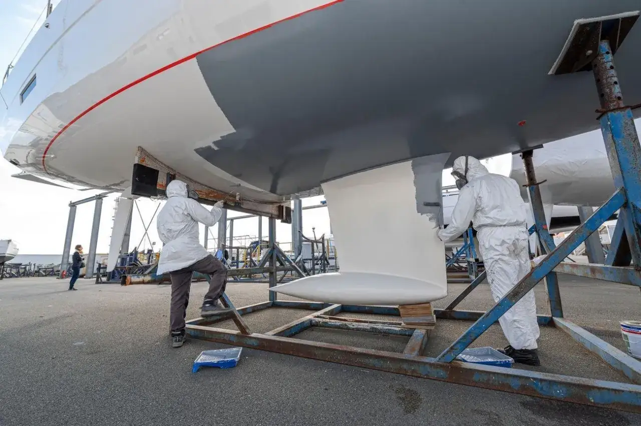 Zwei Arbeiter erneuern das Antifouling eines Bootes. Sie tragen Schutzanz&uuml;ge und malen den Rumpf mit grauer Farbe.