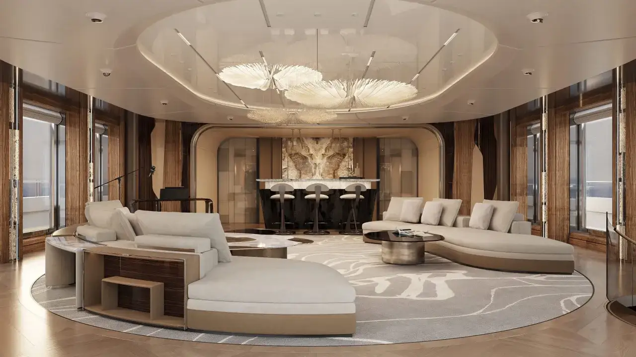 Luxuriöser Salon einer Segelyacht innen mit beigen Sofas, einem runden Couchtisch, einer Bar mit Hockern und auffälligen Deckenleuchten.