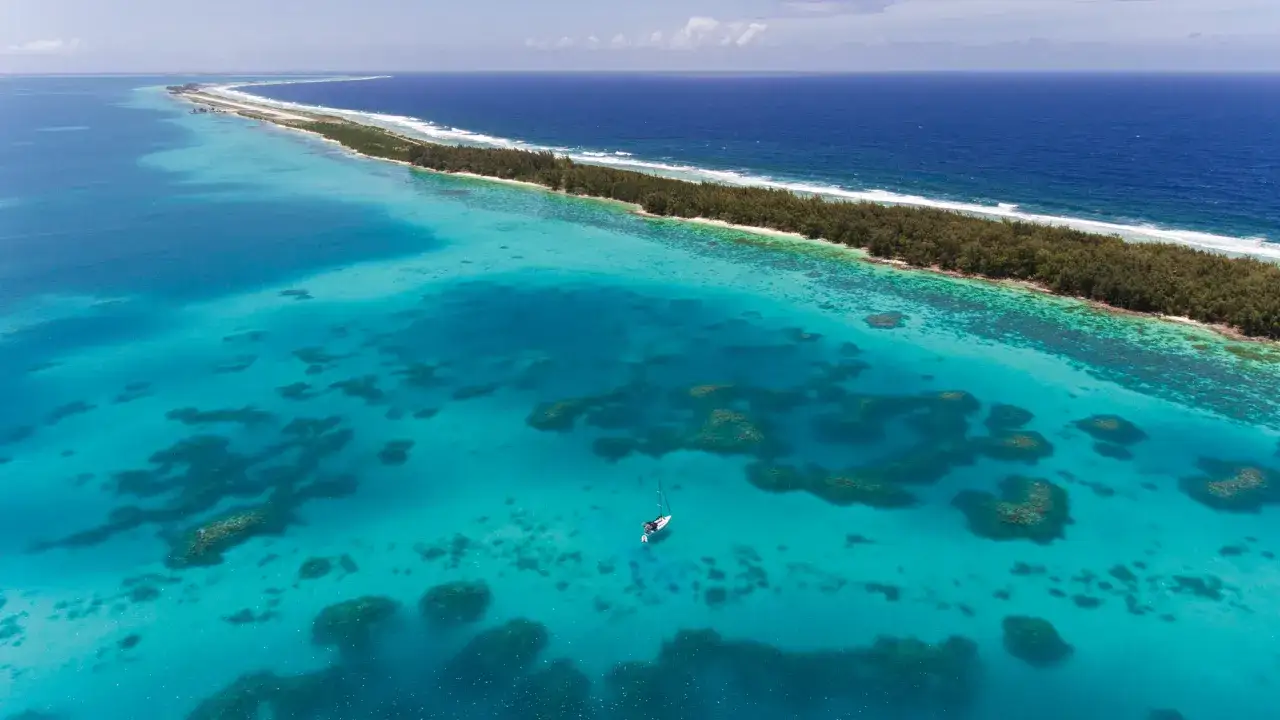 Traumhafte Lagunen und eine einsame Segelyacht vor den Gambier Inseln. Türkises Wasser, Korallenriffe und eine schmale Inselkette.
