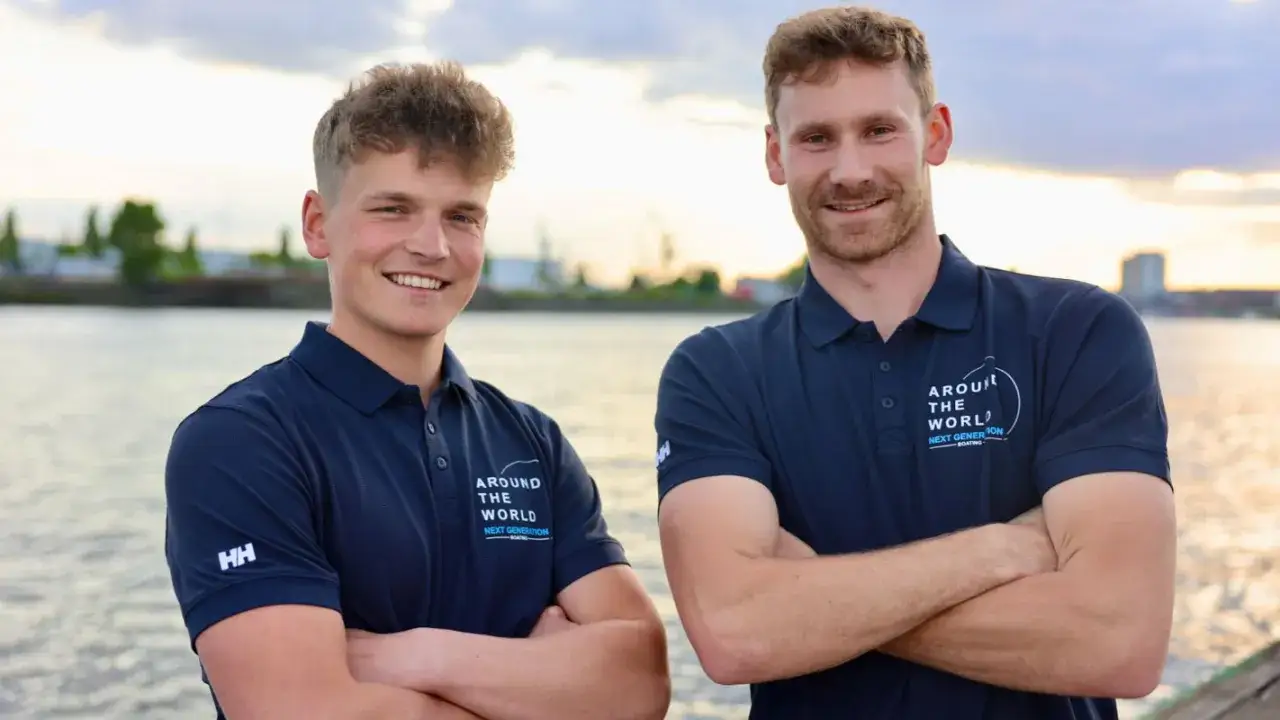 Melwin Fink und sein Teamkollege posieren lächelnd am Wasser. Sie tragen marineblaue Poloshirts mit dem Aufdruck