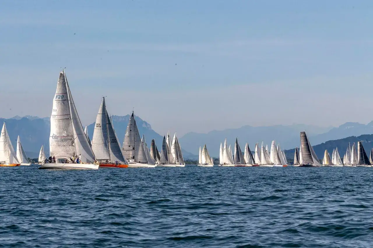 Viele Segelboote k&auml;mpfen bei einer Regatta um die F&uuml;hrung. Die Boote sind auf dem blauen Wasser, mit Bergen im Hintergrund.