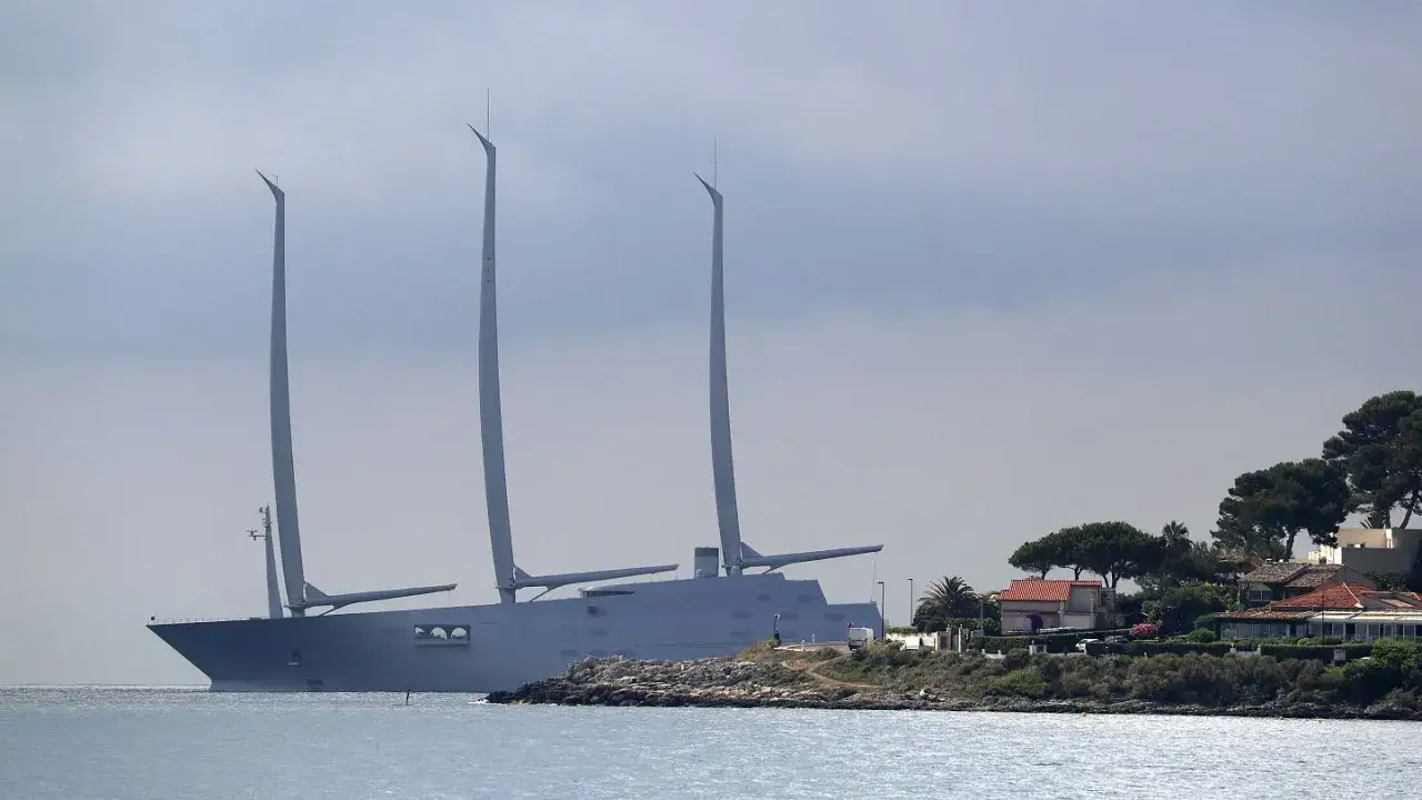 Ein riesiges, graues Segelschiff mit drei hohen Masten liegt vor einer K&uuml;ste mit H&auml;usern und B&auml;umen.