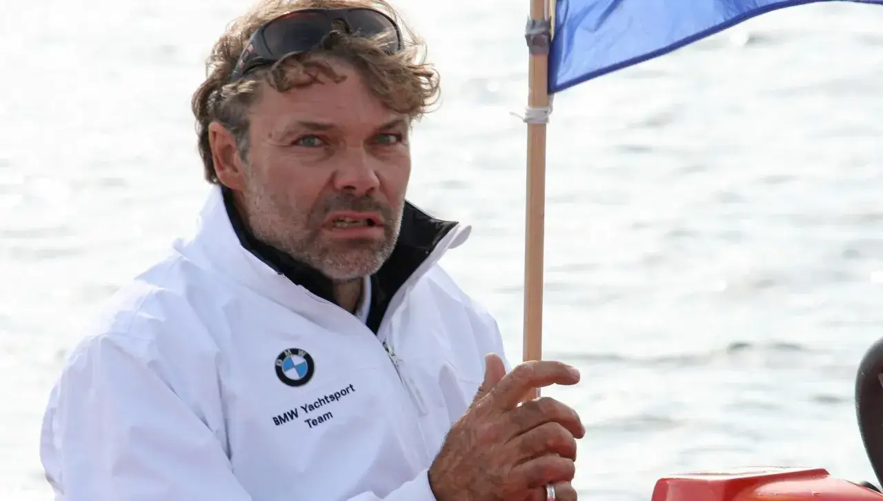Markus Wieser, mit zerzaustem Haar und Sonnenbrille auf dem Kopf, trägt eine weiße BMW Yachtsport Team Jacke. Er hält einen Holzstab mit einer blauen Flagge.
