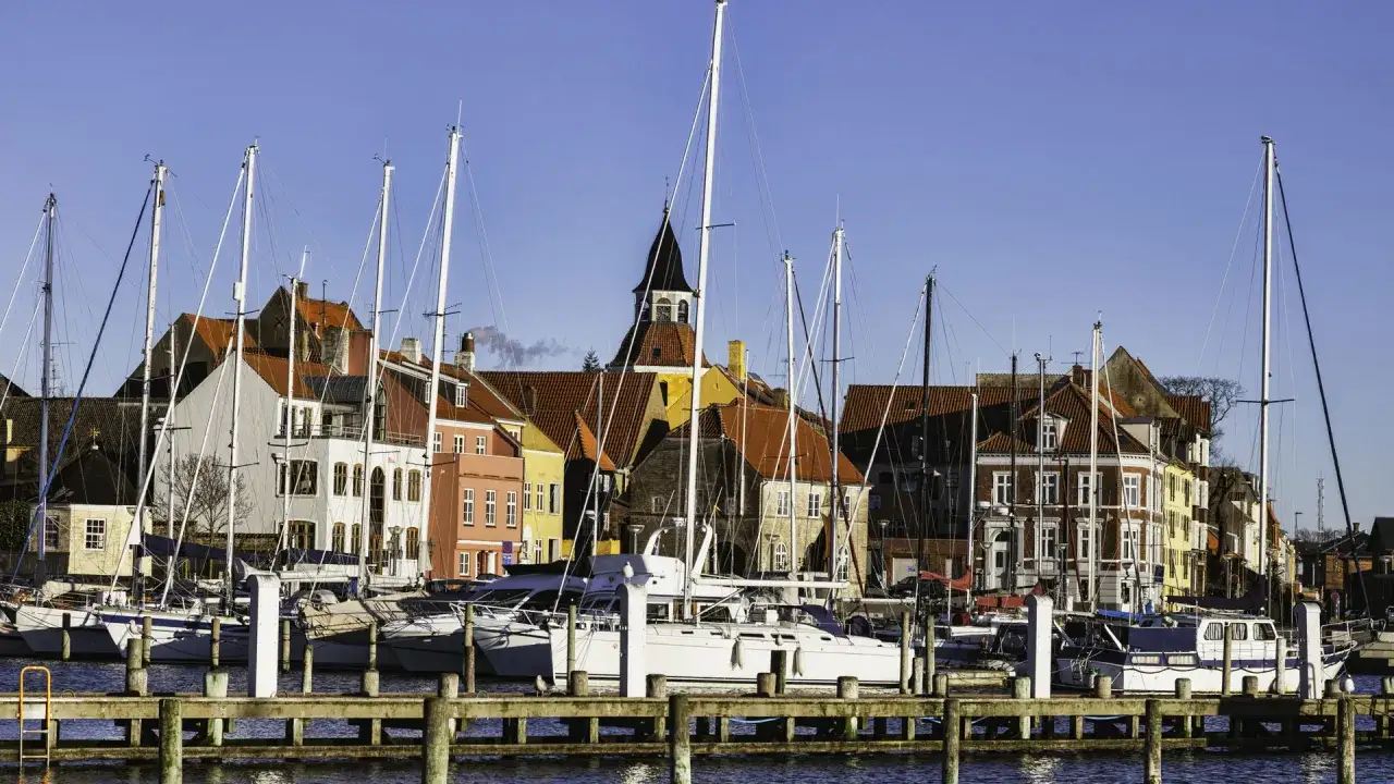 Segelboote im Flensburg Hafen vor bunten H&auml;usern und einem Kirchturm unter blauem Himmel.