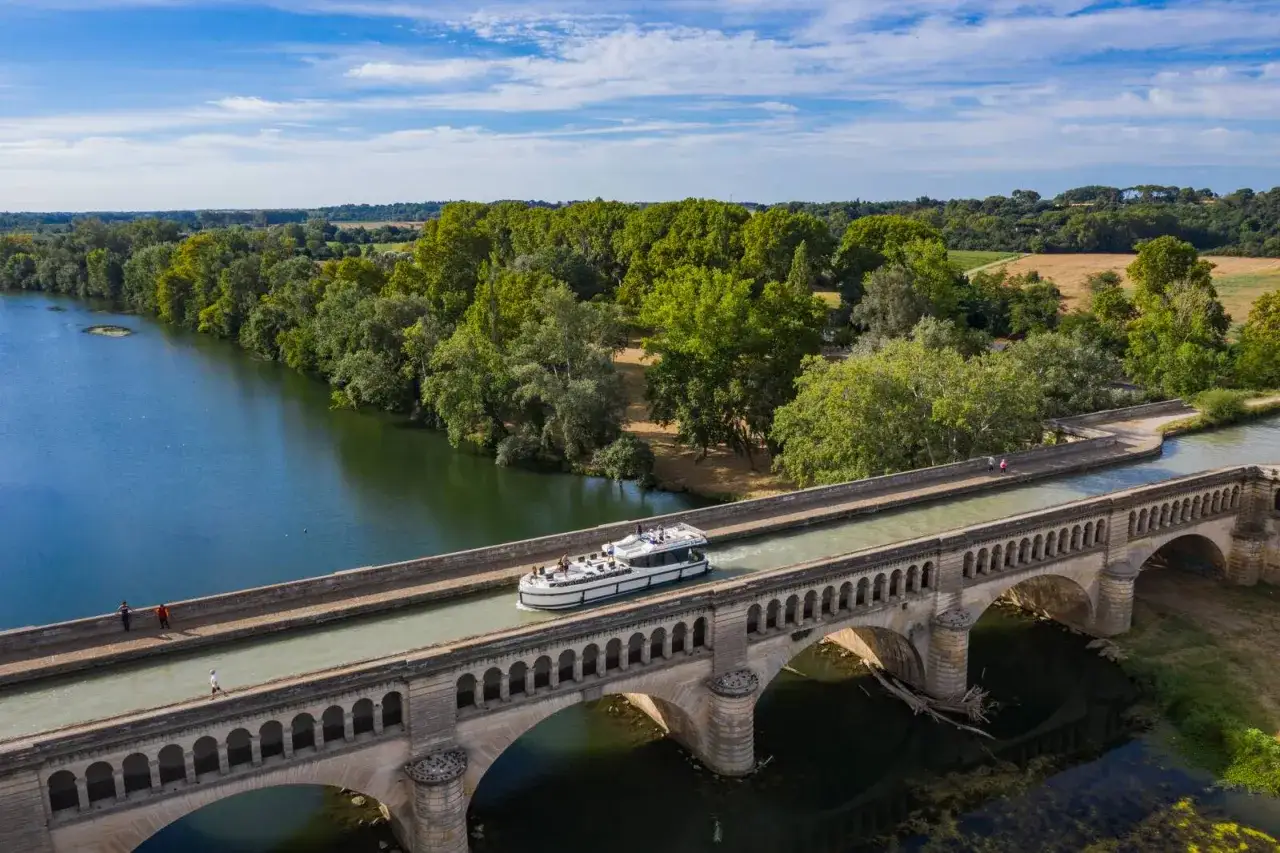 Ein Hausboot f&auml;hrt &uuml;ber eine Br&uuml;cke auf einem Kanal in Frankreich. Gr&uuml;ne B&auml;ume und blauer Himmel im Hintergrund.