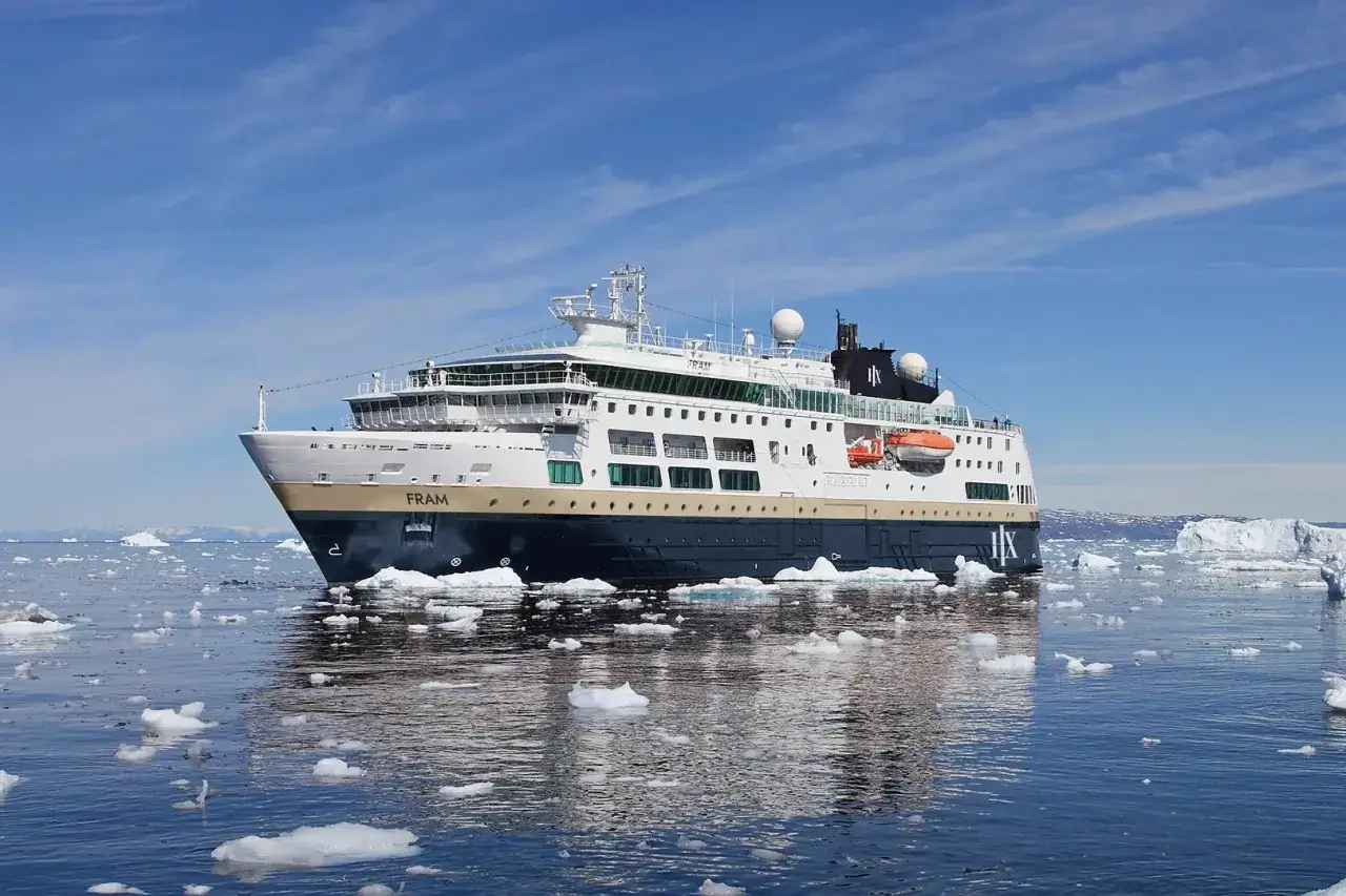 Das Schiff Fram navigiert durch eisige Gewässer, umgeben von kleinen Eisbergen unter einem strahlend blauen Himmel.