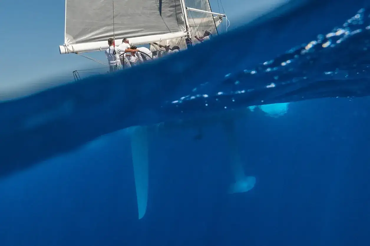Unterwasseraufnahme des segelboot rumpf mit Crew an Deck. Kiel und Ruderblatt ragen in das klare blaue Wasser.