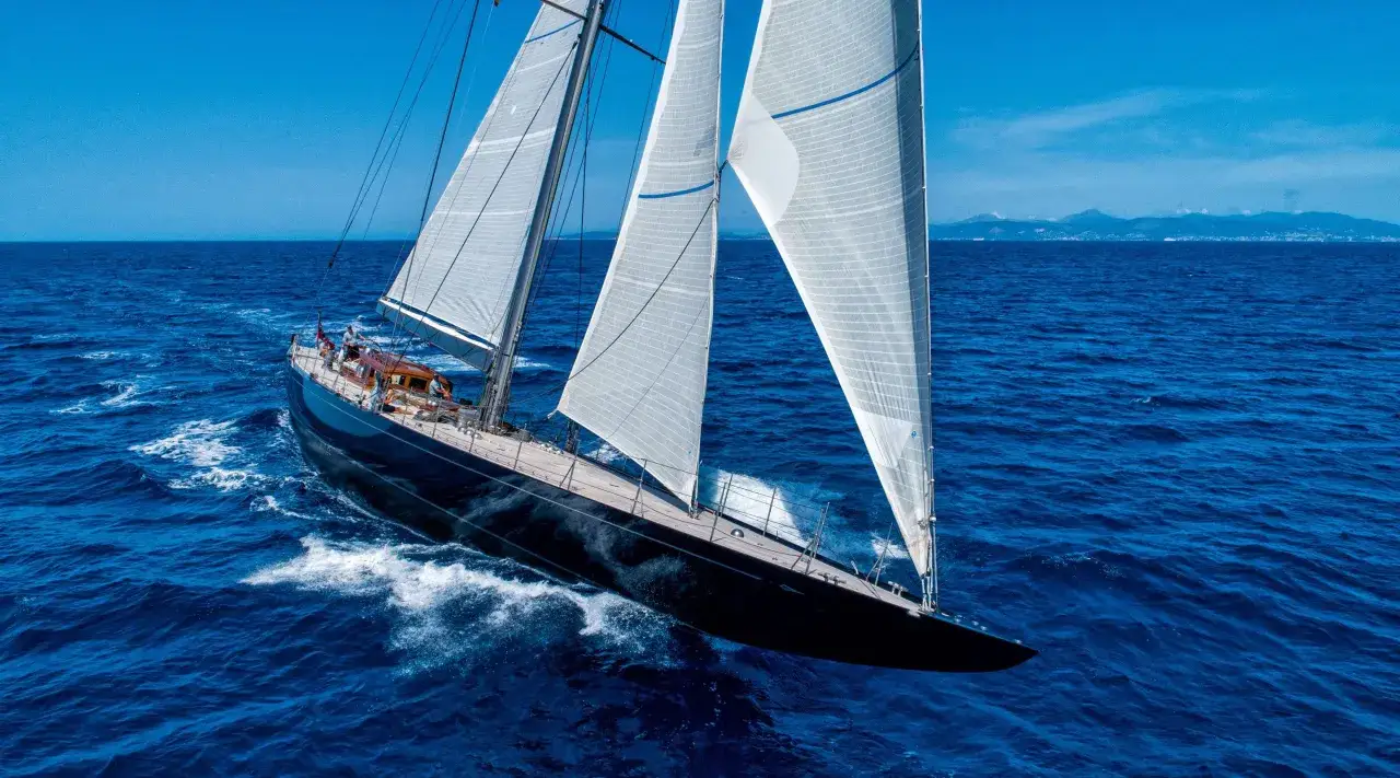 Segelyacht der j class gleitet auf tiefblauem Meer, wei&szlig;e Segel im Wind, im Hintergrund eine K&uuml;stenlinie.