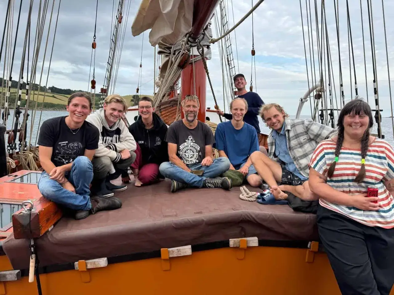 Fröhliche Crew genießt das Segeln in Zeeland. Acht Freunde sitzen auf dem Deck eines Segelschiffs, umgeben von Takelage und mit Blick auf eine grüne Küste.