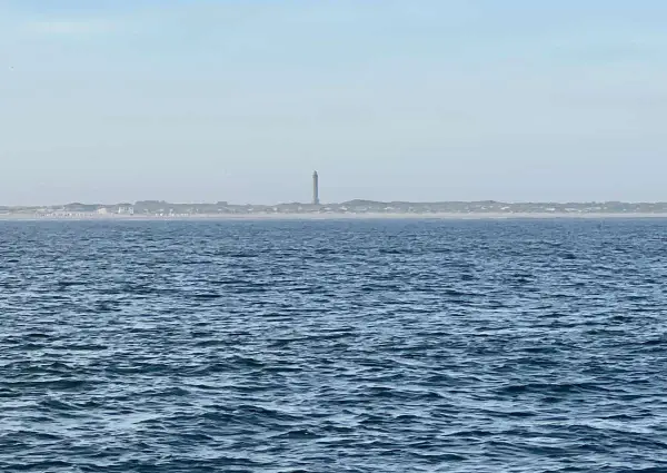 Der Leuchtturm Norderney ragt über die Dünenlandschaft am Horizont. Das weite Meer liegt davor.