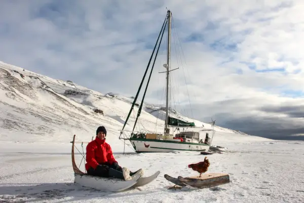 Guirec Soudée sitzt in einem Schlitten vor seinem Segelboot in einer verschneiten Landschaft. Eine Henne steht auf einem Holzbrett daneben.