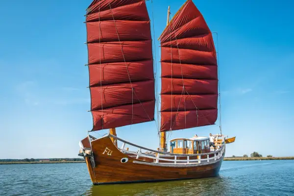 Ein hölzernes Dschunke Schiff mit roten Segeln gleitet auf ruhigem Wasser unter blauem Himmel.