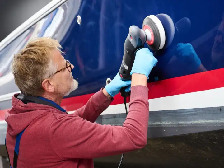 Mann mit Brille poliert mit Maschine den blauen Rumpf eines Bootes.