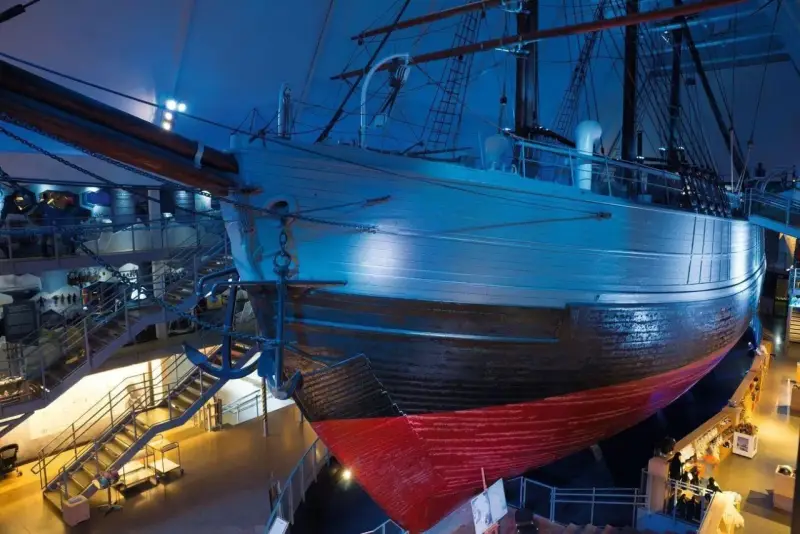 Ein historisches **fram schiff** mit Anker, umgeben von Treppen und Ausstellungsbereichen, in blauem Licht.