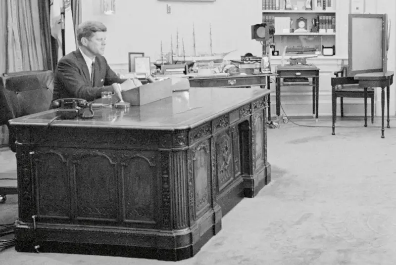 John F. Kennedy am Schreibtisch im Oval Office. Hinter ihm ein Modellschiff, das an die HMS Resolute erinnert, die dem Schreibtisch seinen Namen gab.