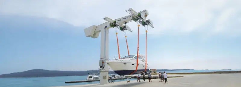 Ein großer Kran hebt ein weißes Boot aus dem Wasser. Mehrere Personen beobachten den Vorgang.