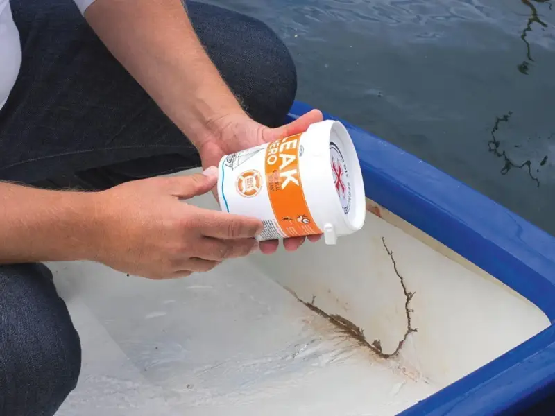 Person hält Dose mit Dichtmittel, um ein Boot abzudichten. Riss im Bootsrumpf sichtbar.