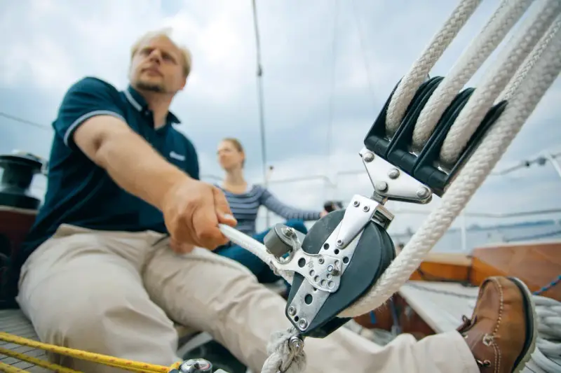 Mann zieht mit kräftiger Hand das Tauwerk am Segel. Ein Mann und eine Frau auf einem Segelboot, die das Tauwerk am Segel bedienen.